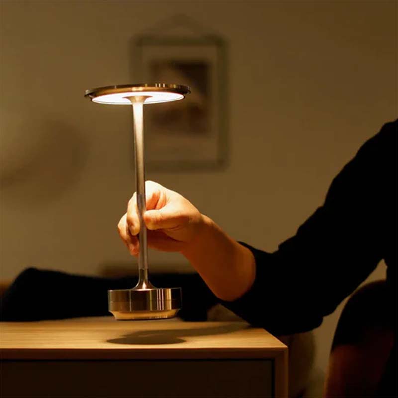 Man holding portable metal table lamp over wooden surface against blurred background.