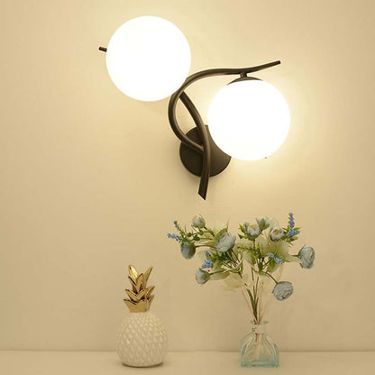 Wall-mounted light fixture with two round lights, a small pineapple-shaped vase, and a clear vase with flowers on a beige surface.