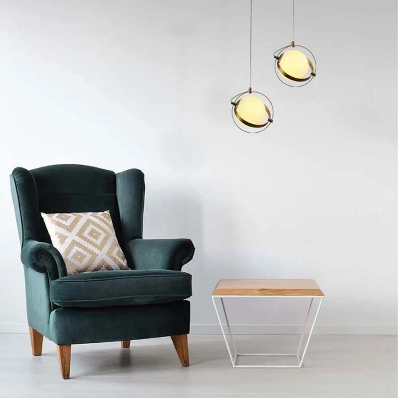 A golden framed globe pendant with gold trim hangs over a green armchair with a patterned pillow and a small wooden table on light gray floors.