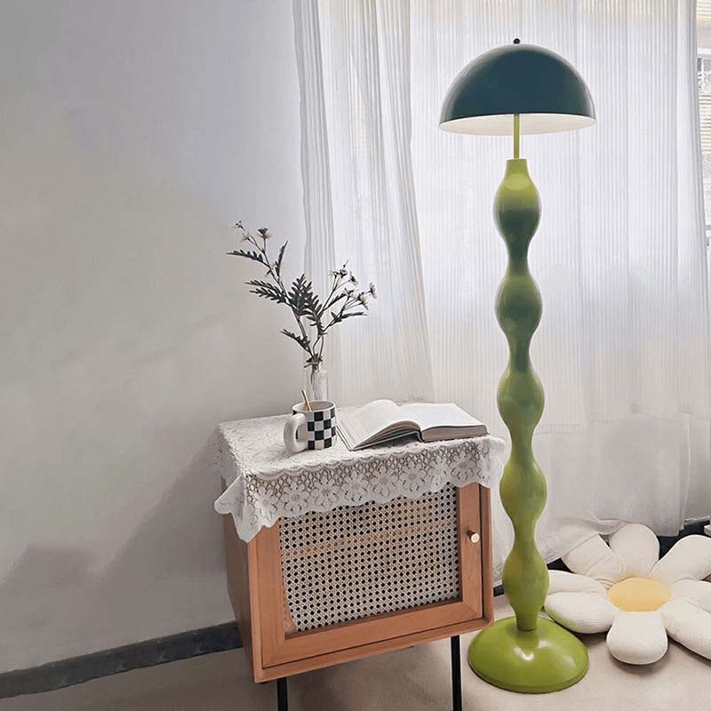 Green floor lamp next to a side table with a vase and book in a room with white curtains.