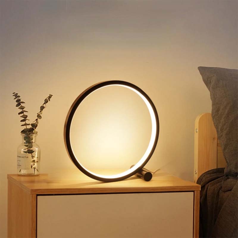 Round table lamp on a wooden nightstand with a vase of eucalyptus in a bedroom setting.