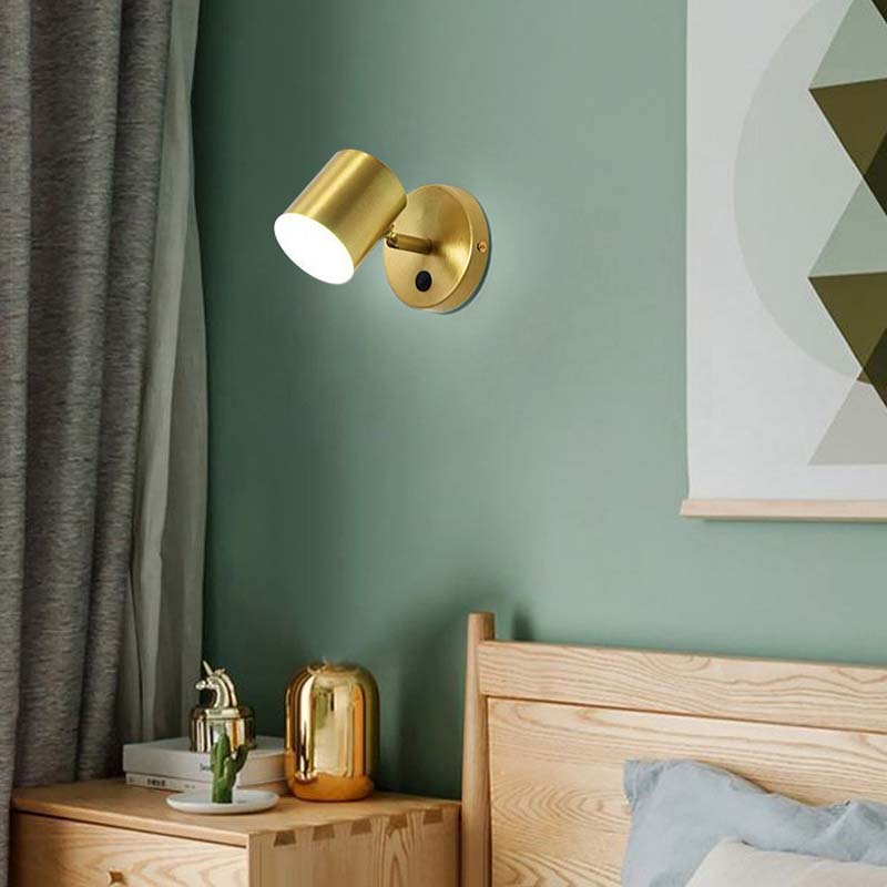 Gold wall sconce on a green wall with a wooden bed and side table in the foreground.