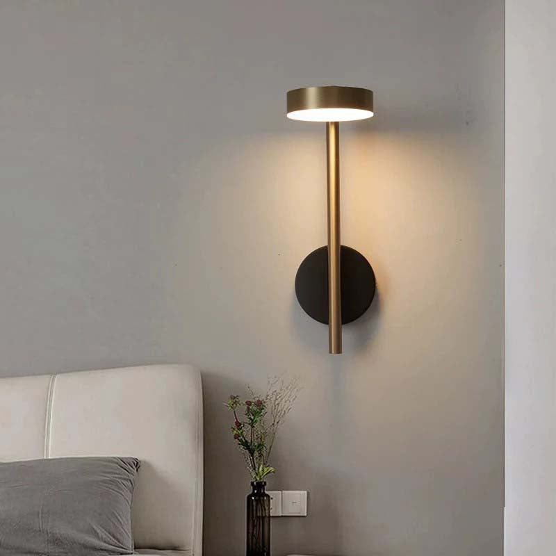 Modern wall sconce light fixture on a gray wall with a bed and vase in the background.