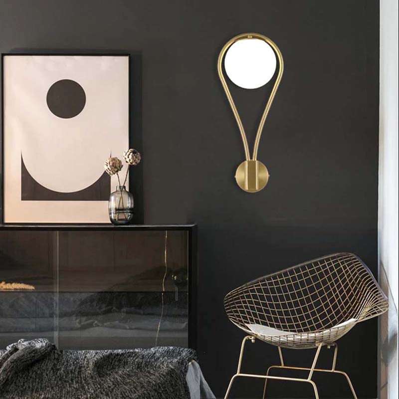Modern interior design with a gold wall sconce, abstract art, and wire chair against a black wall.