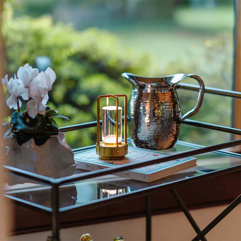 Decorative lantern on a table with a pitcher and flowers, blurred greenery in the background