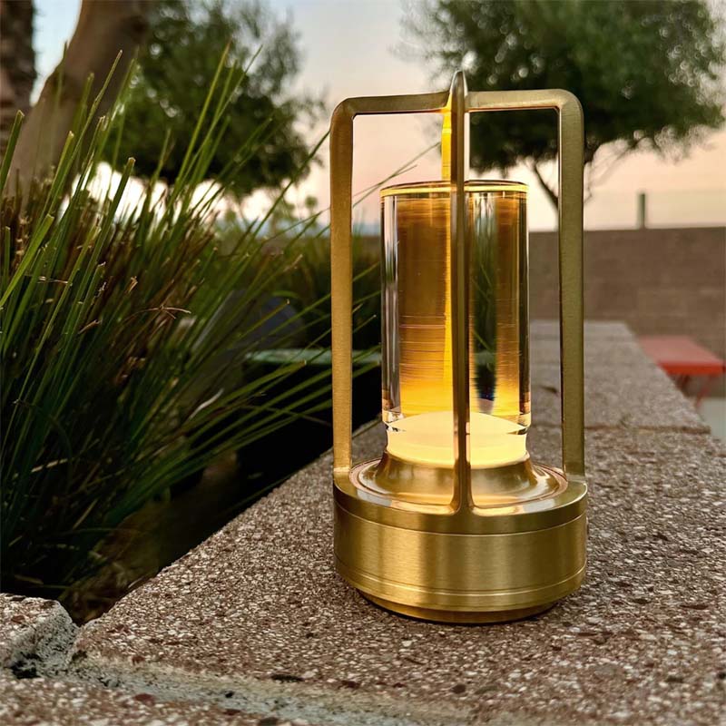 Gold outdoor lantern on a patio with plants and a bench in the background