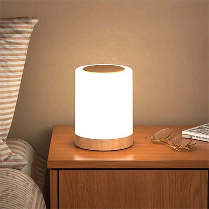 Table lamp on a wooden nightstand with a warm glow, next to a bed and glasses.