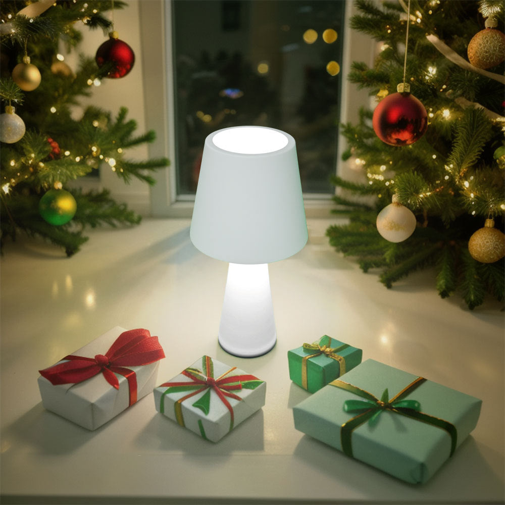 White table lamp on a surface with Christmas decorations including trees and wrapped gifts.