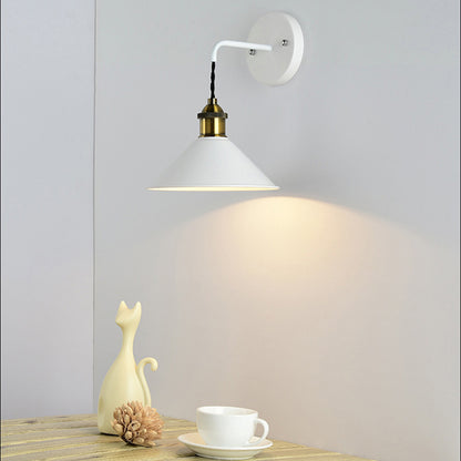 Wall-mounted lamp with a white shade and gold fixture on a light gray wall, with a decorative cat figurine and cup on a wooden surface in the foreground.
