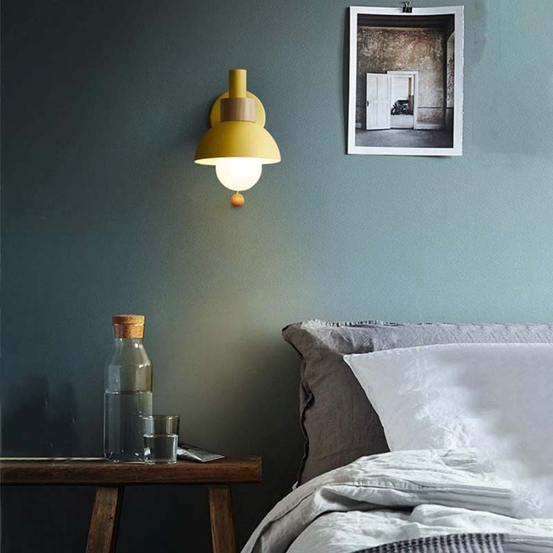 Yellow wall lamp on a dark green wall above a wooden nightstand with a bottle and glass.