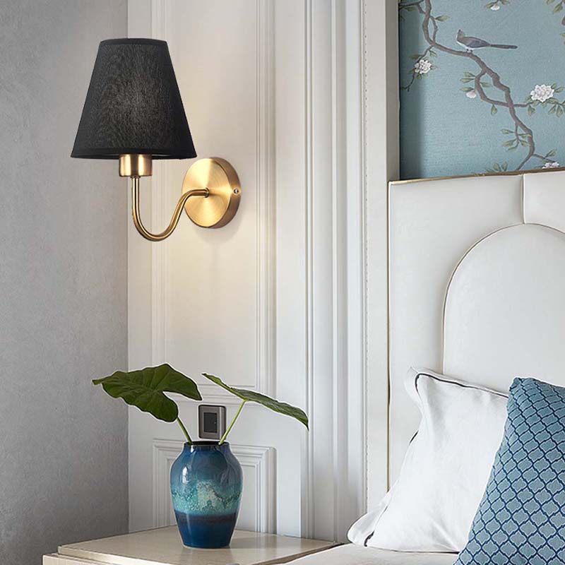 Decorative wall sconce with black shade and gold fixture on a white wall, next to a vase with green leaves and a framed artwork.