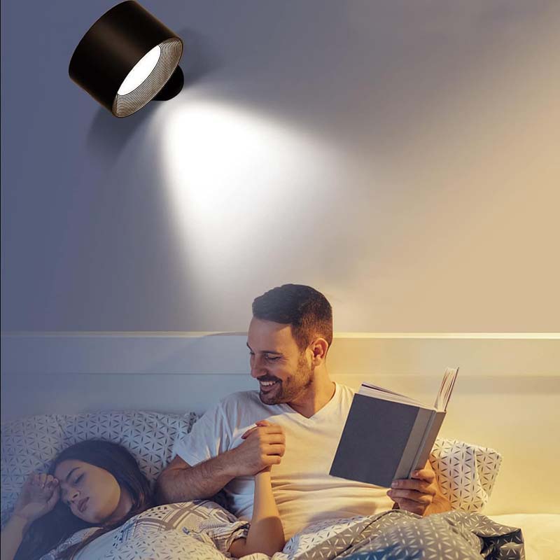 Man reading a book to a woman in bed under a wall-mounted light fixture.