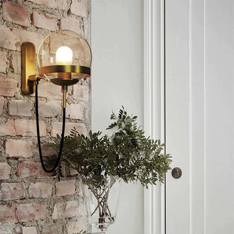 Wall-mounted brass light fixture on a brick wall with a plant in the foreground.