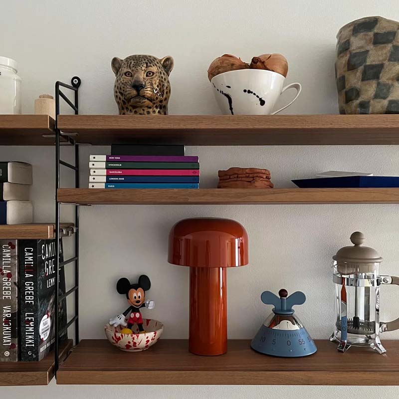 The Rechargeable Mushroom LED Table Lamp sits on a shelf in a stylish interior composition featuring wooden hanging shelves against a white wall, displaying books, ceramic mugs, figurines, and decorative items.
