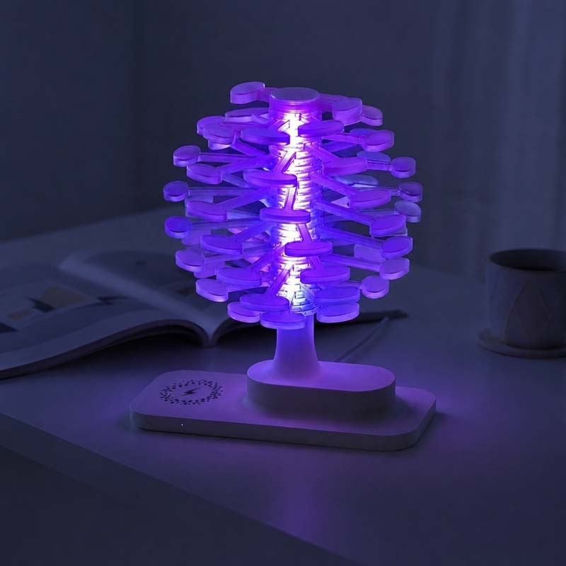 Spiral Tree LED Desk Lamp with purple glow inside, placed on a white base on a table next to an open book and a cup in a dimly lit room.