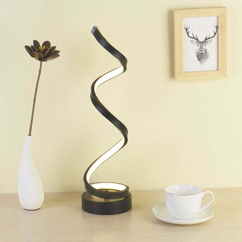 A modern spiral twist table lamp with a black frame and glowing white light, styled on a wooden table next to a white vase with a flower and a white teacup on a saucer, against a beige wall.