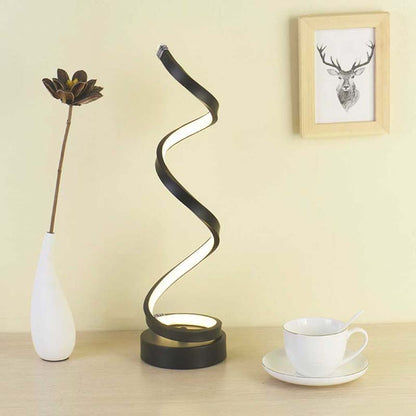 A modern spiral twist table lamp with a black frame and glowing white light, styled on a wooden table next to a white vase with a flower and a white teacup on a saucer, against a beige wall.