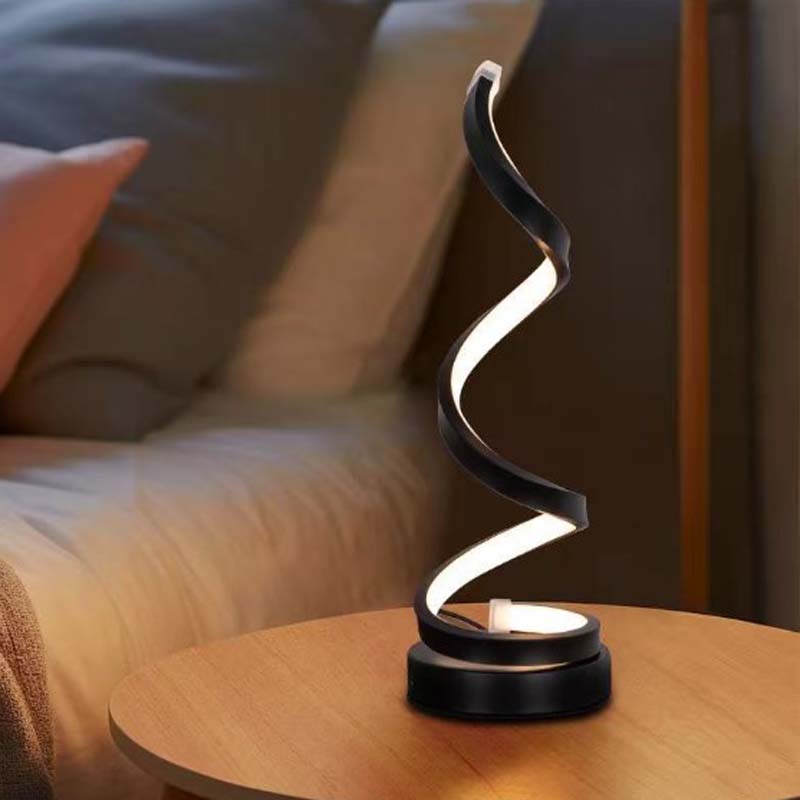 A modern spiral-shaped table lamp with a black frame and glowing white light, placed on a round wooden side table in a softly lit bedroom setting.