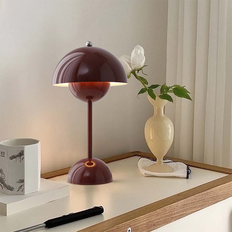 Table lamp with a maroon shade on a wooden surface next to a vase with a flower.