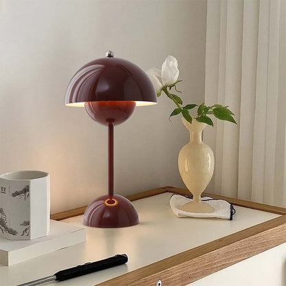 Table lamp with a maroon shade on a wooden surface next to a vase with a flower.