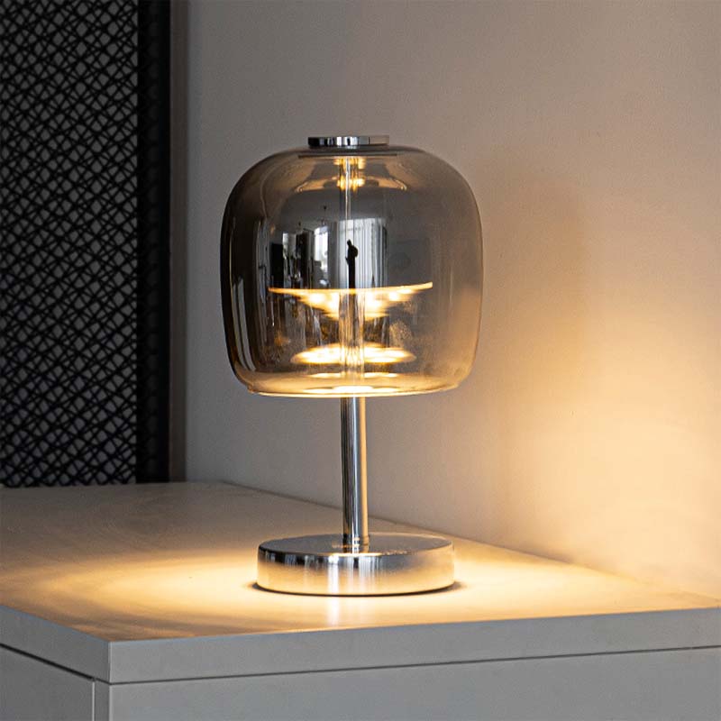 A modern table lamp with a clear glass dome shade and a polished silver metal base, illuminated with a warm yellow light on a tabletop.