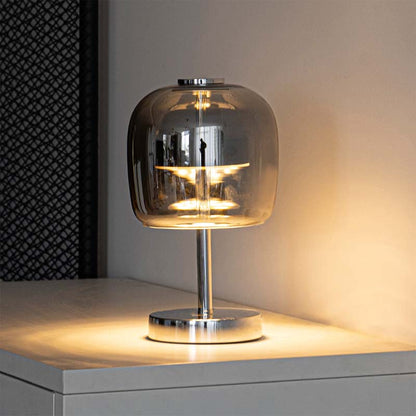 A modern table lamp with a clear glass dome shade and a polished silver metal base, illuminated with a warm yellow light on a tabletop.