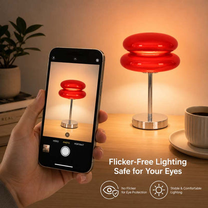 A man holds a smartphone with an image of a Retro Sandwich Glass Table Lamp on its screen, along with a similar lamp lit on a wooden table in a warm room. The caption on the image reads, "Flicker-free, safe for your eyes."