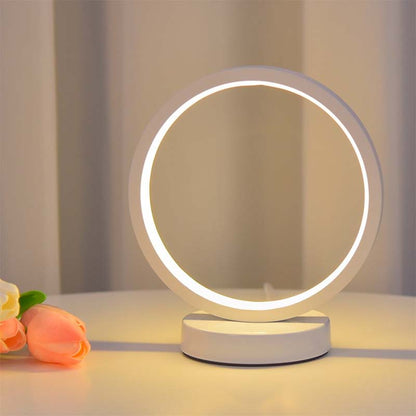 Minimalist LED ring table lamp with soft warm light on a white surface, with a flower nearby