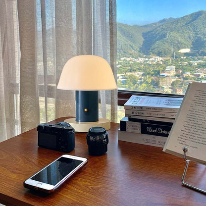 A table with a Nordic Mushroom LED Table Lamp, a camera, a phone and books, with a picturesque view outside the window.