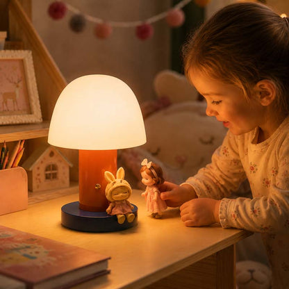 A little girl with brown hair sits at a wooden table, looking at a small doll, and the warm light of the Nordic Mushroom LED Table Lamp illuminates the scene.