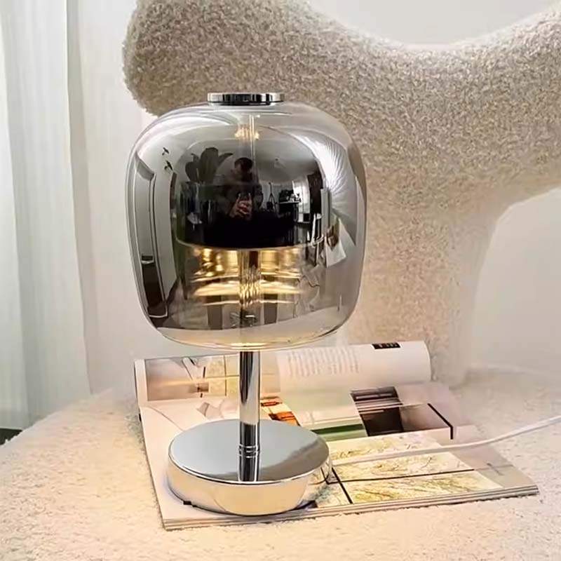 A modern table lamp with a highly polished chrome dome-shaped shade and circular base, resting on an open magazine in front of a textured cream chair.