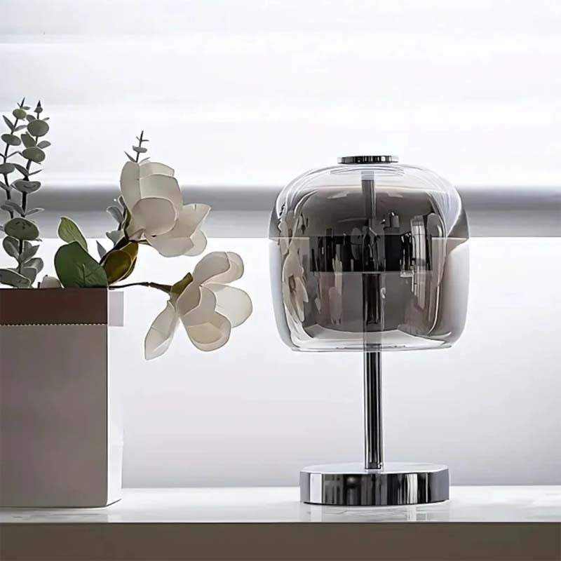 Modern table lamp with transparent glass shade on a white surface with plants in the background