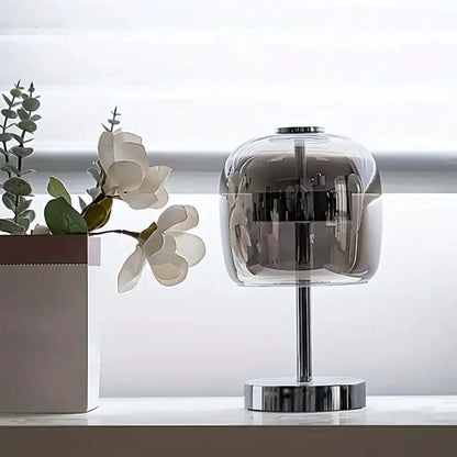 Modern table lamp with transparent glass shade on a white surface with plants in the background