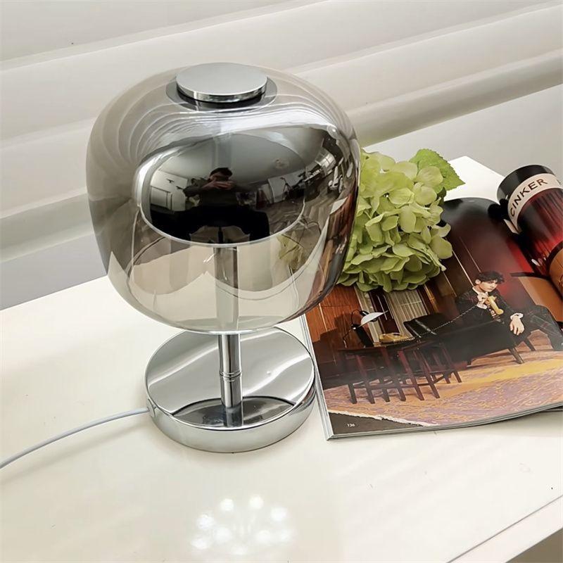 A modern polished chrome table lamp with a spherical mirrored shade and circular base, shown on a white tabletop next to a magazine and green hydrangea.