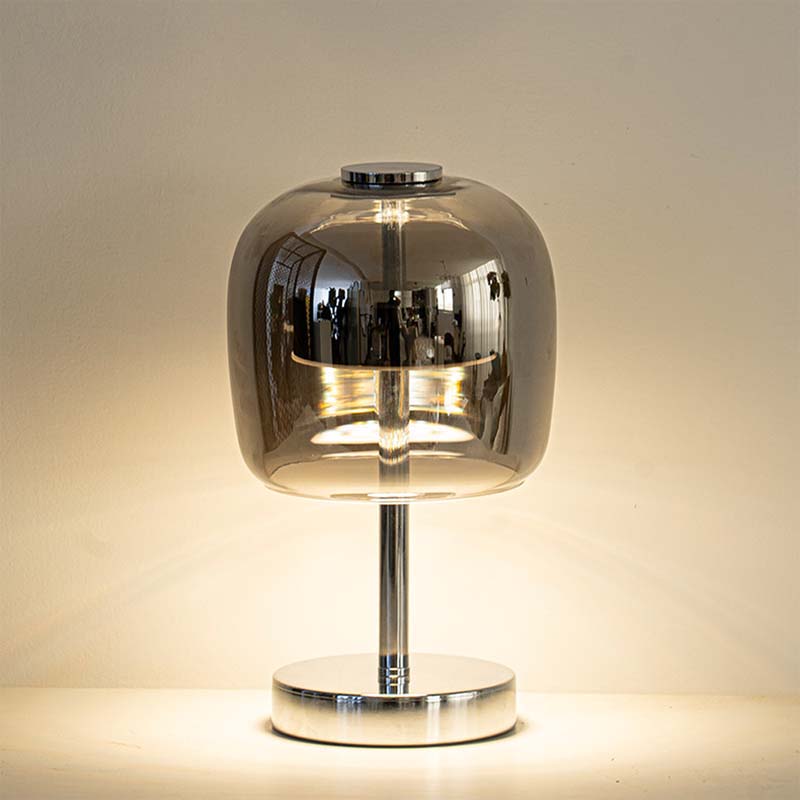 A modern table lamp with a rounded, dome-shaped smoked glass shade on a polished chrome stand and circular base, illuminated against a warm beige background.