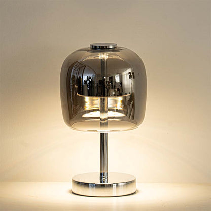 A modern table lamp with a rounded, dome-shaped smoked glass shade on a polished chrome stand and circular base, illuminated against a warm beige background.