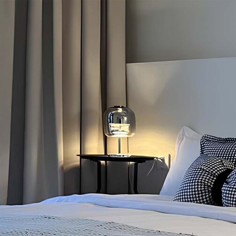 Premium Glass LED Table Lamp sits on a black bedside table next to a bed with white bedding and black and white pillows in a softly lit bedroom.