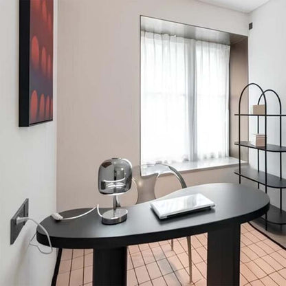 Modern minimalist home office featuring a black oval desk, Premium Glass LED Table Lamp, white chair, and black metal shelving against a beige wall.