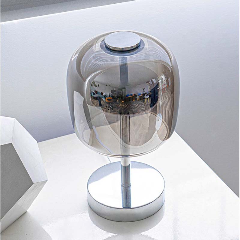 A modern table lamp with a transparent smoky dome shade and a polished silver metal base, shown on a white surface.