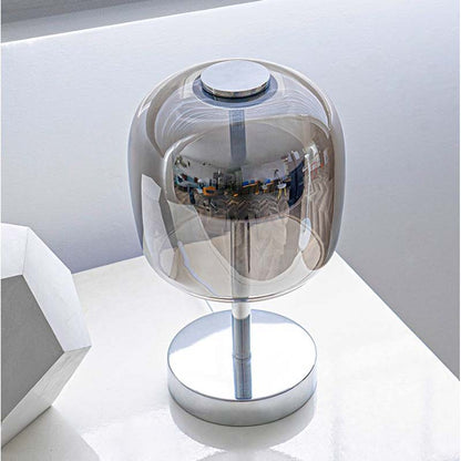 A modern table lamp with a transparent smoky dome shade and a polished silver metal base, shown on a white surface.