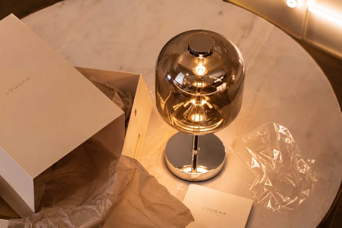 A lit modern table lamp with a spherical amber-tinted glass shade and polished metallic base, shown beside its branded box and tissue paper on a round marble table.
