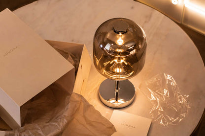 A lit modern table lamp with a spherical amber-tinted glass shade and polished metallic base, shown beside its branded box and tissue paper on a round marble table.