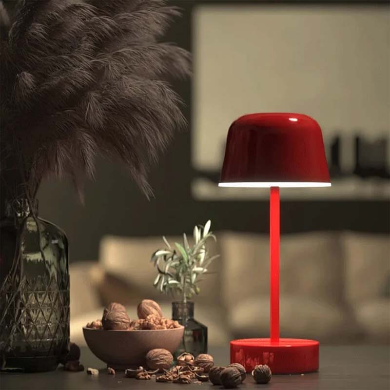 A red Rechargeable Cordless Touch LED Table Lamp sits on a surface with decorative objects in a blurred interior.