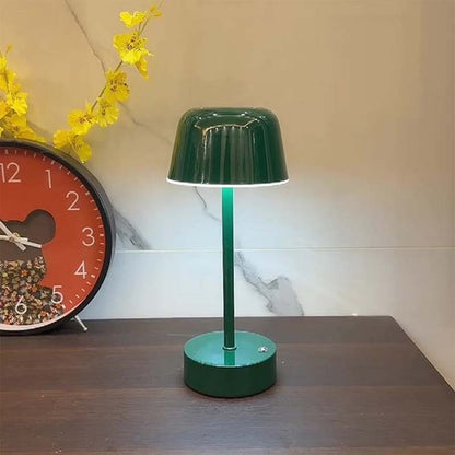 The Rechargeable Cordless Touch LED Table Lamp sits on a dark wooden tabletop against a light wall. A round orange clock and yellow flowers are visible in the background.