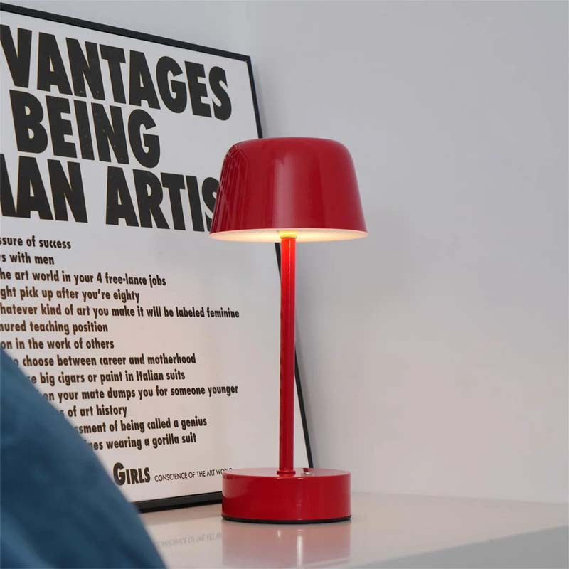 A Rechargeable Cordless Touch LED Table Lamp stands on a white surface, with a poster with black text on a white background hanging behind it.