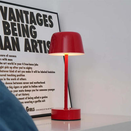 A Rechargeable Cordless Touch LED Table Lamp stands on a white surface, with a poster with black text on a white background hanging behind it.