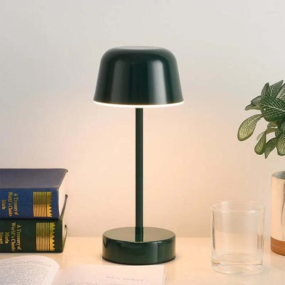 A black Rechargeable Cordless Touch LED Table Lamp sits on a table, with books and a plant in the background.
