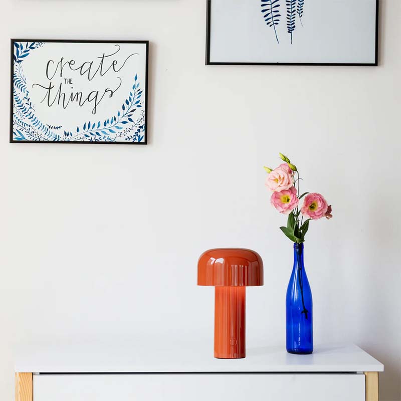 The Rechargeable Mushroom LED Table Lamp sits on a shelf in a stylish interior composition with a white wall and two framed paintings.