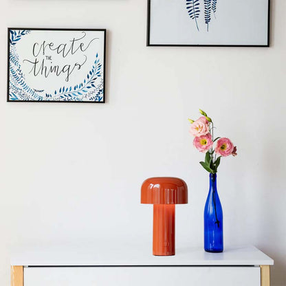 The Rechargeable Mushroom LED Table Lamp sits on a shelf in a stylish interior composition with a white wall and two framed paintings.