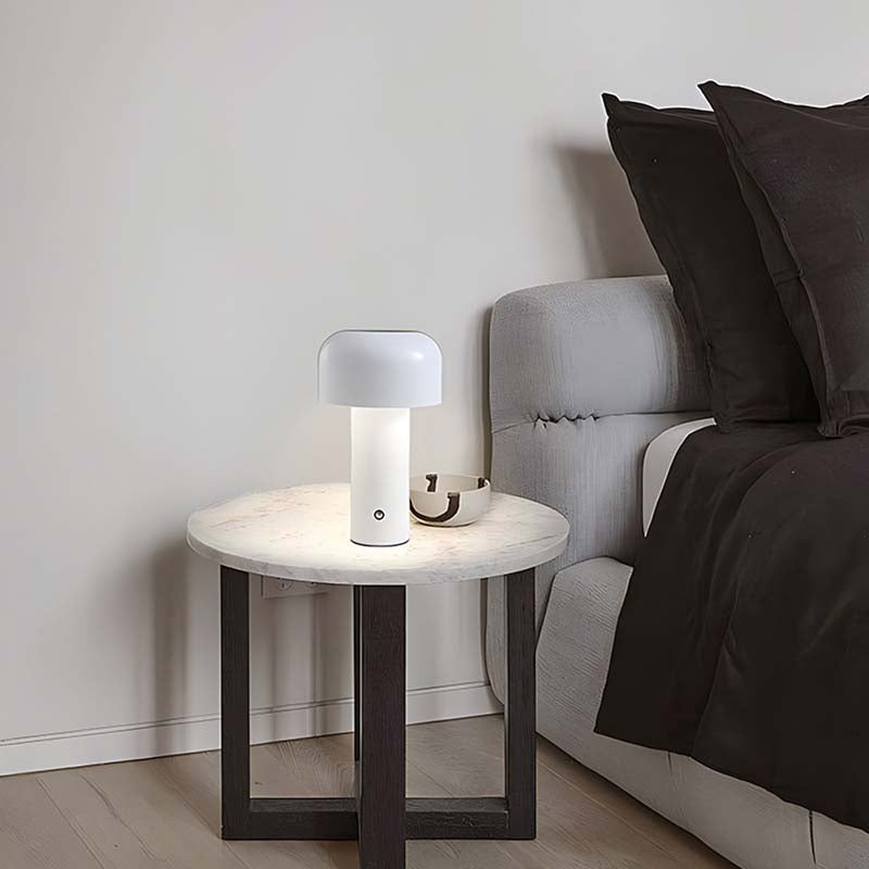 The Rechargeable Mushroom LED Table Lamp sits on a round nightstand with a marble top next to a bed dressed in dark linens and pillows.